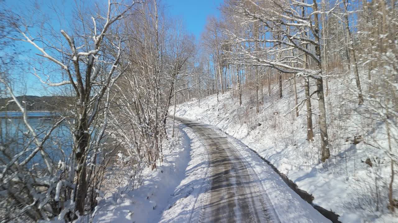 Drone flying along winding winter road covered in snow next to frozen Teåkersjön lake shoreline in rural Dalskog Mellerud Sweden