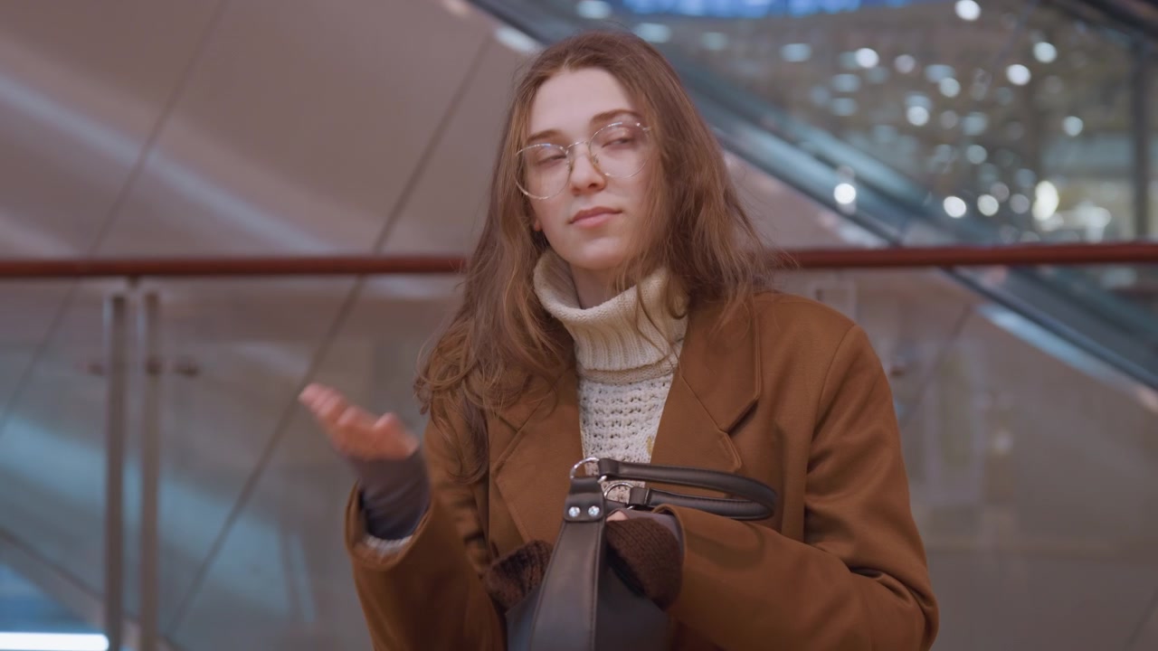 Donna cerca nella borsa su una panchina, apre una borsa di pelle e ne controlla il contenuto con espressione curiosa, luci serali della stazione e ringhiera di vetro sullo sfondo, gesti e piccoli movimenti rivelano sorpresa