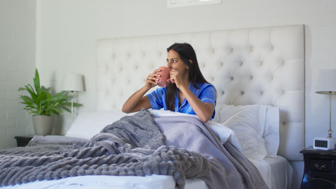 Donna adulta con vestiti di raso blu seduta a letto che sorseggia da una tazza rosa, sente un rumore e si volta.