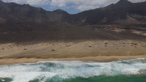 Fantastische-Luftaufnahme,-Die-Die-Wunderschönen-Berge-Enthüllt,-Die-Den-Strand-Von-Cofete-Umgeben,-Und-Die-Wellen,-Die-Sich-Am-Ufer-Brechen