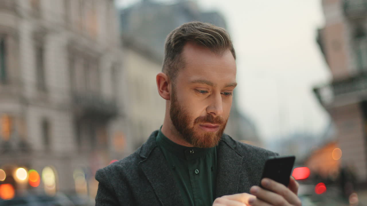 Close-up view of caucasian businessman with a beard texting on the phone in the street with city lights of the background