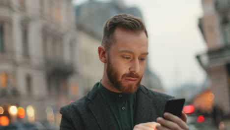 vista de cerca del hombre de negocios caucásico con barba enviando mensajes de texto por teléfono en la calle con luces de la ciudad de fondo