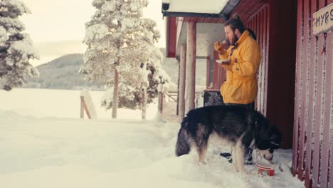 man and pet dog alaskan malamute eating meal outside the cabin on a winter day