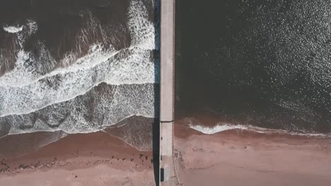 Aerial-birds-eye-view-drone-footage-of-a-waves-crashing-onto-the-beach-against-a-pier,-Roker-Beach,-Sunderland,-UK