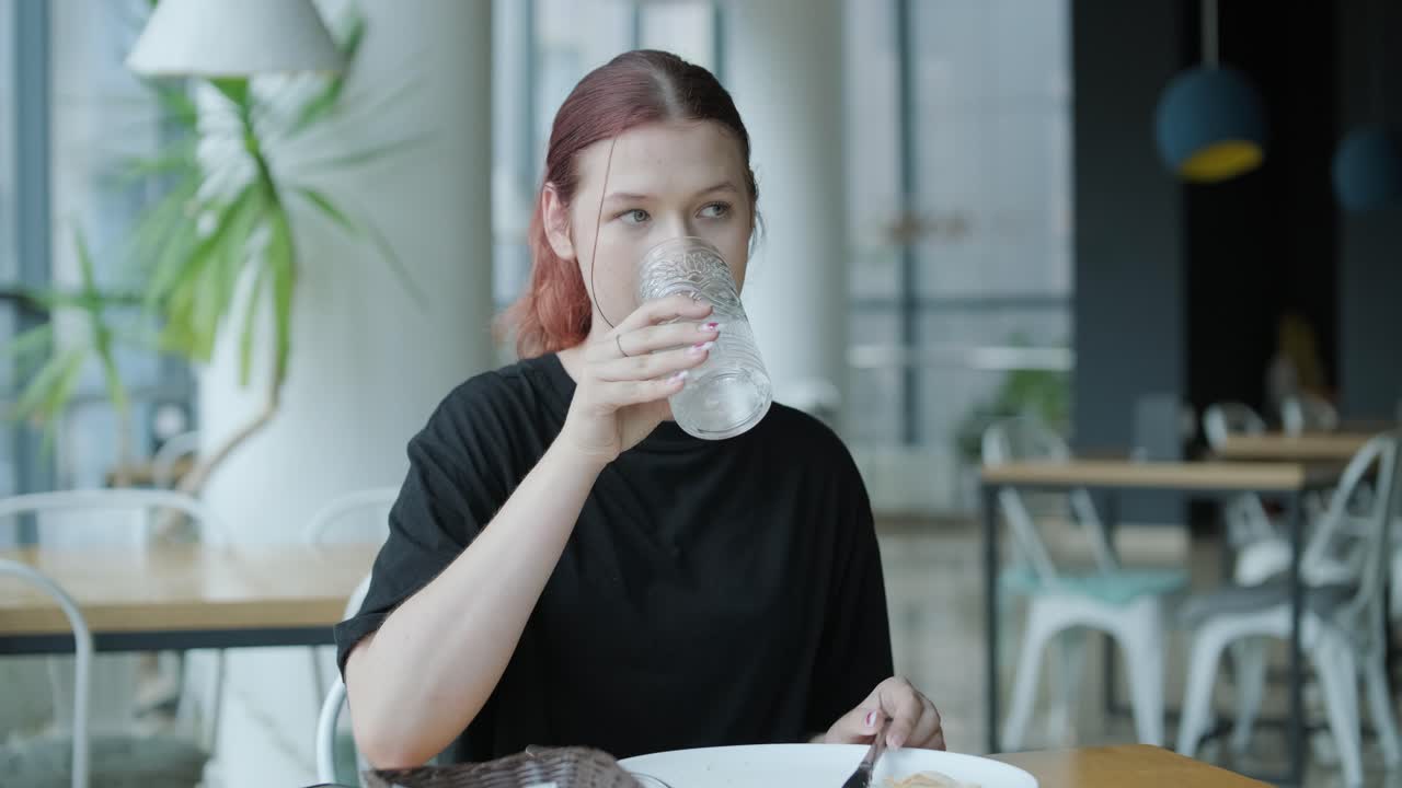 젊은 여성이 큰 창문, 자연빛, 도시 전망을 가진 현대적인 레스토랑에서 나무 테이블에서 신선한 러드를 먹고 있습니다. 편안한 식사 장면