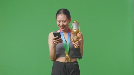 asian woman with a gold medal holding a gold trophy and using smartphone on green screen background in the studio