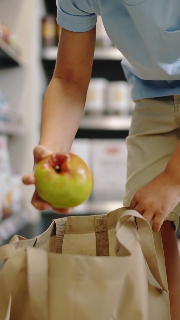 Vertical video: Leaning boy in blue T reaching in bag at grocery aisle selecting green apple to eat