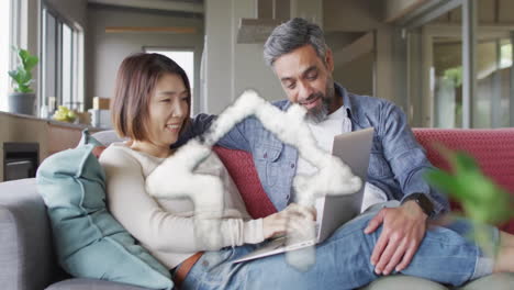 animation of house shape over diverse couple sitting on sofa using laptop at home