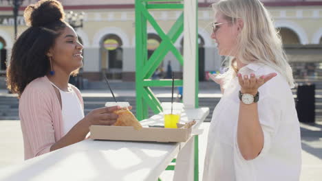 -Female-Friends-Chatting-And-Laughing-While-Sharing-Pizza-And-Drinking,-Standing-At-An-Outdoor-Table-In-The-Street-1