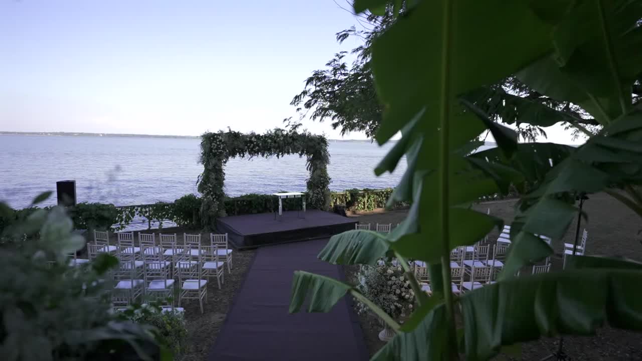 La ripresa con svelamento lento passa dalle foglie tropicali in primo piano all'elegante allestimento di una cerimonia nuziale all'aperto, mostrando arco floreale, palco, sedie bianche e uno sfondo di acqua calma prima dell'arrivo degli ospiti