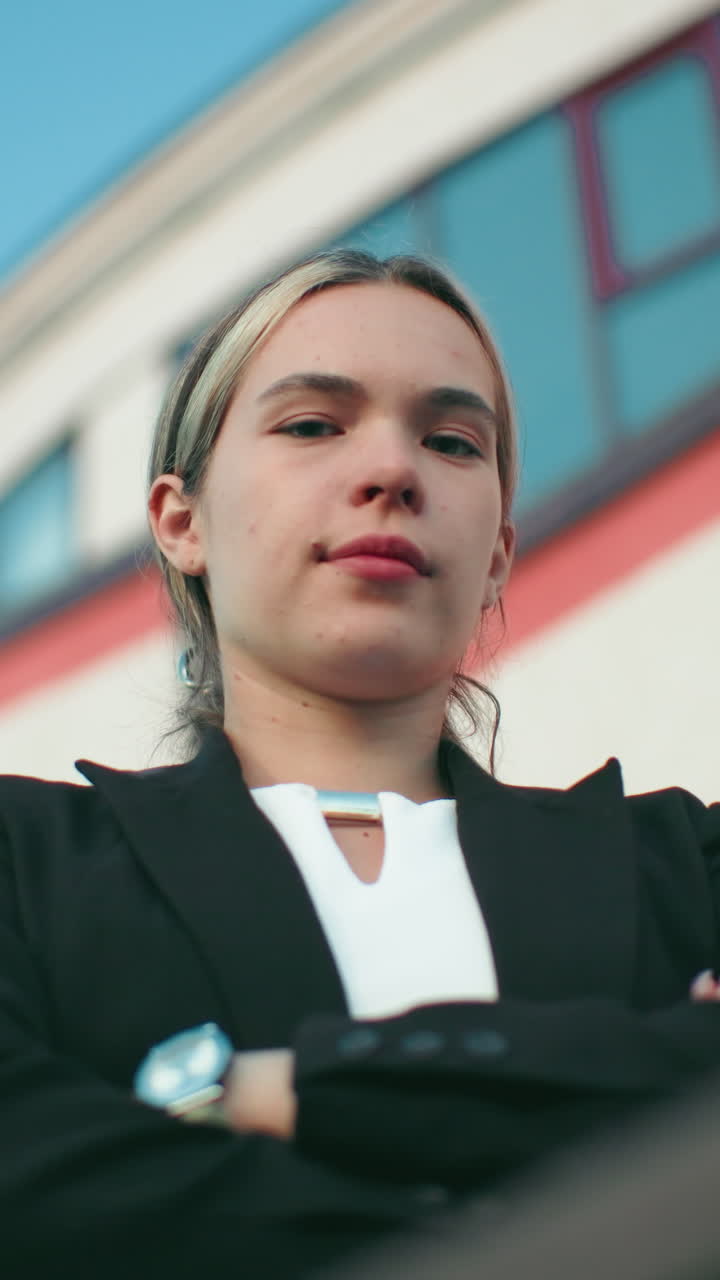 Young manager in black blazer folds hands confidently while standing in front of organization building with red and white walls and visible security camera