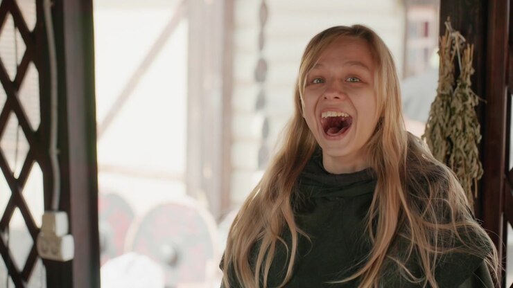 Young woman stands in wooden doorway laughing with mouth open, eyes closed, head tilted, and hair swept by motion; warm daylight backlights face and hair, creating soft glow, relaxed mood, and