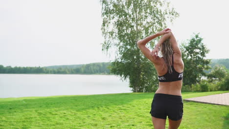 A-slender-and-beautiful-woman-in-sports-clothes-with-an-open-press-is-preparing-to-start-training-on-the-lawn-near-her-home-stretching-her-arms-and-shoulders-with-warming-up-exercises