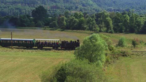 An aerial drone captures a historic steam train full of passengers. Tourists enjoy a scenic journey through a rural landscape with hay bales, a perfect summer holiday excursion