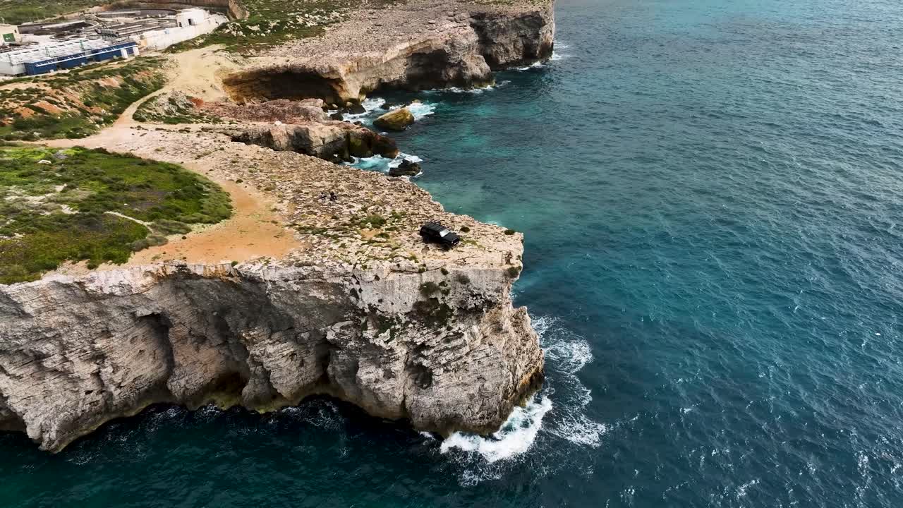 Aerial footage captures a black off-road vehicle perched dramatically on the jagged limestone cliffs of Ras Tanit, surrounded by turquoise Mediterranean waters and wild coastal scenery.