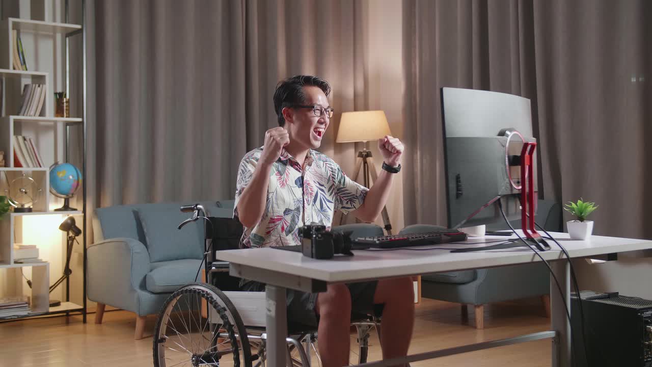 hombre asiático en silla de ruedas celebrando el éxito de la edición del video por un escritorio junto a la cámara en casa