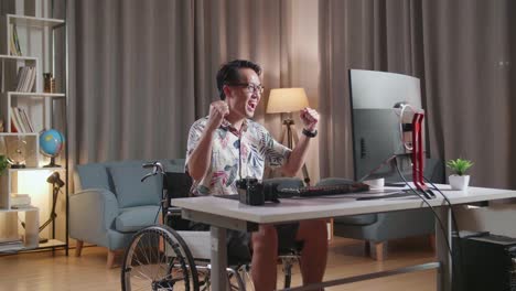 hombre asiático en silla de ruedas celebrando el éxito de la edición del video por un escritorio junto a la cámara en casa