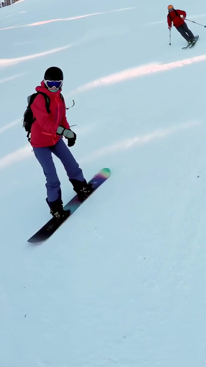 Video verticale: Scarico della seggiovia mentre uno snowboarder con giacca rosa scia verso l'obiettivo sulla pista per rallentare.