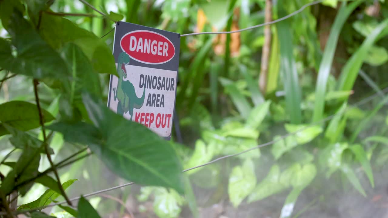 A red and white warning sign reading 'Danger Dinosaur Area Keep Out' stands among dense green foliage, with subtle camera movement and natural daylight