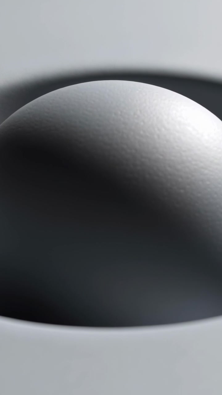 Video verticale: La telecamera si allontana, rivelando una sfera grigio opaco in un piatto, mostrando uno studio di luce soffusa.