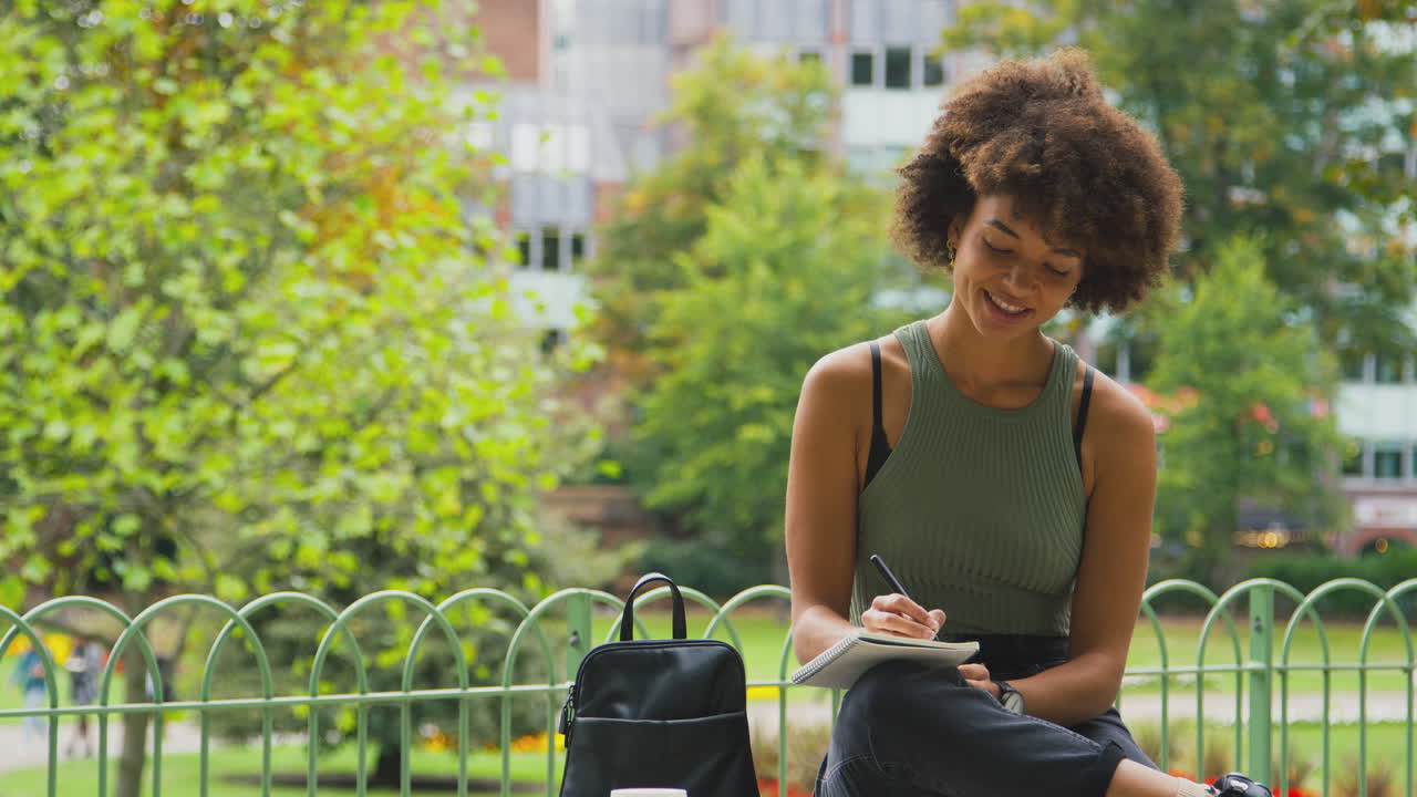 Jeune femme en extérieur dans un parc assise sur un banc travaillant et écrivant dans un carnet