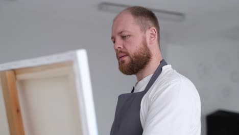 a bearded man in an apron looking at a canvas on an easel
