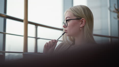 student thoughtfully taking notes while gazing outside. she holds a pen in her hand, lost in deep thought, wearing glasses and a cozy sweater in a calm, bright study space near a window