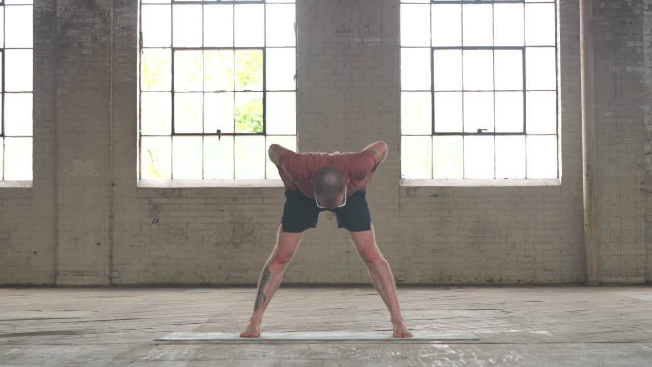 Man in an industrial warehouse practicing yoga doing a wide legged forward fold.