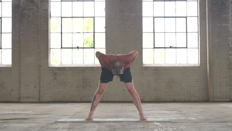 man in an industrial warehouse practicing yoga doing a wide legged forward fold.