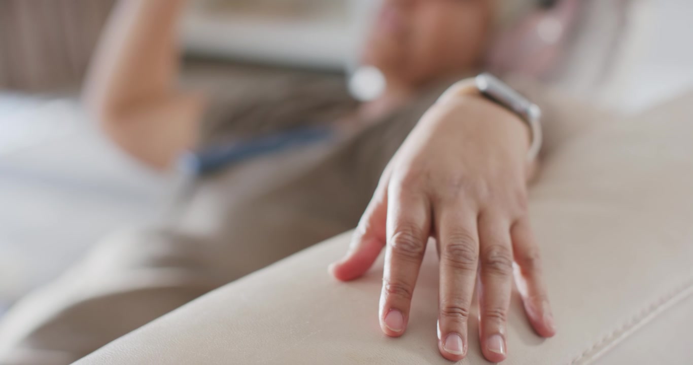 Woman resting right wrist on sofa armrest at home, smartwatch on wrist, left hand checking device