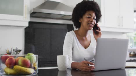 Afroamerikanische-Frau,-Die-In-Der-Küche-Mit-Dem-Smartphone-Spricht-Und-Einen-Laptop-Benutzt,-Während-Sie-Von-Zu-Hause-Aus-Arbeitet