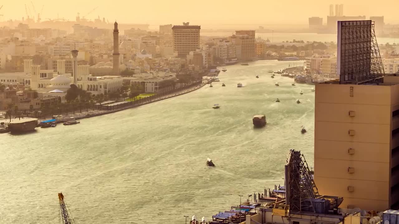 Riprese in time-lapse di una vivace città fluviale al tramonto, che proietta un caldo bagliore dorato sulla scena. Numerose imbarcazioni navigano il canale, riflettendo il trasporto attivo e il commercio