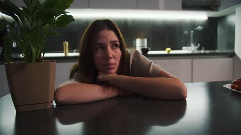 A-sad,-thoughtful-brunette-girl-in-a-beige-T-shirt-leans-on-the-table-and-thinks-about-something-sad-near-a-houseplant-on-a-black-table-in-the-kitchen-in-a-modern-apartment