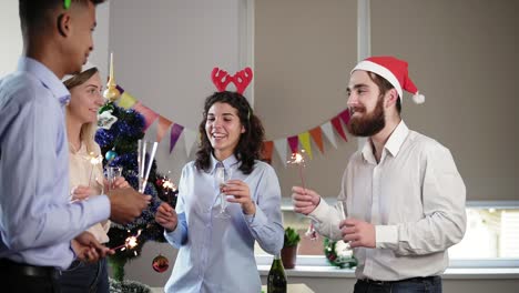 multi-ethnic-group-of-happy-office-workers-holding-Bengal-lights-and-dancing-wearing-christmas-hats-and-deer-headband-and