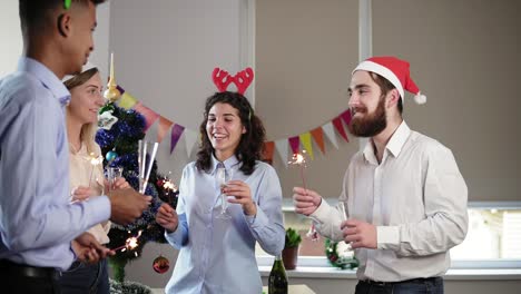 grupo multiétnico de trabajadores de oficina felices sosteniendo luces de bengala y bailando usando sombreros de navidad y diadema de ciervo y