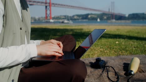 person working on laptop in park by the river with bridge in background