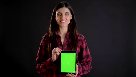 retrato de una joven caucásica de cabello largo sonriendo mostrando la pantalla verde de una tableta vertical sobre un fondo negro.