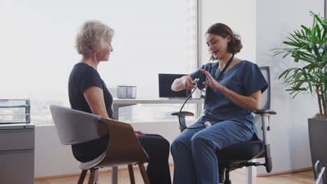 female doctor wearing scrubs in office listening to senior female patients chest using stethoscope