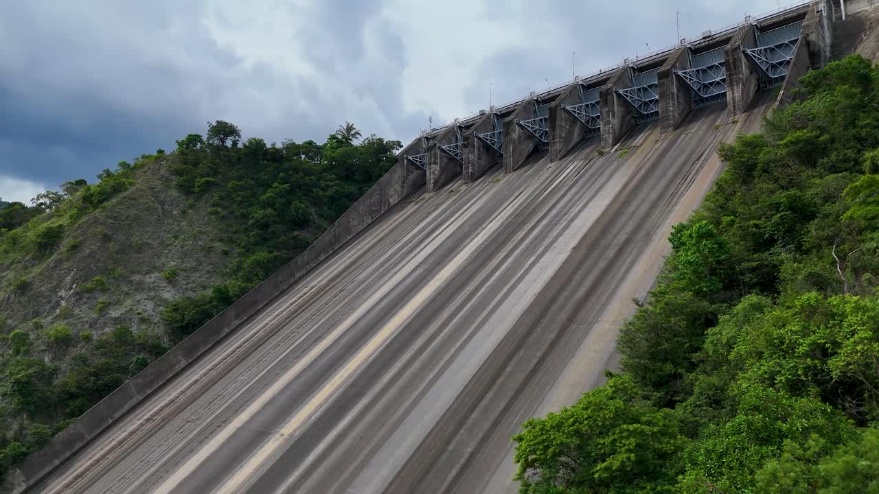 Massive concrete dam structure beside a lush green hillside under a cloudy sky