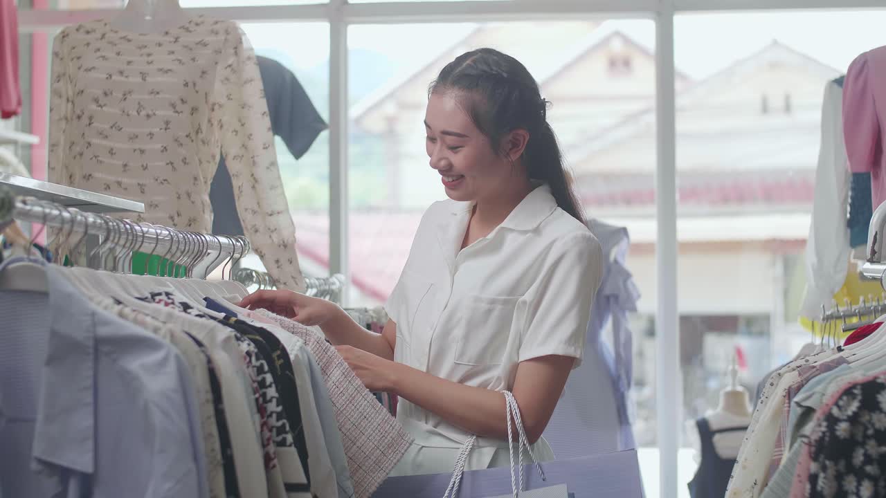 쇼핑 가방을 들고 있는 젊은 아시아 여성은 부티크에서 쇼핑하는 동안 거에 옷을 선택하는 것을 즐긴다