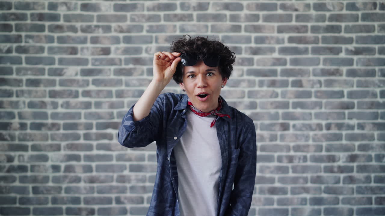 Woman wearing sunglasses and a bandana, surprised expression in front of a brick wall