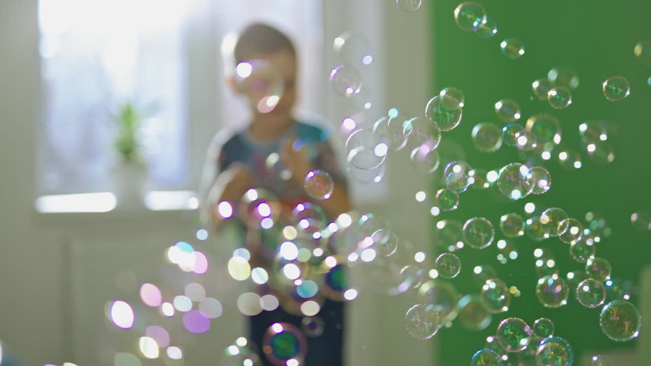 Lots of round soap bubbles in the room. Beautiful colorful bubbles flying in the air indoors and little boy catches bright balls happily. Close-up.