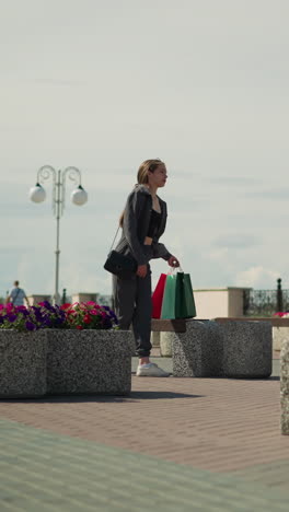dama de ropa gris camina hacia un banco al aire libre, llevando bolsas de compras de colores, se acerca al banco cerca de camas de flores, se sienta, se ve a dos personas caminando en el fondo borroso