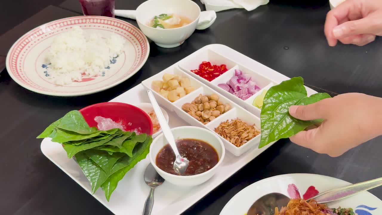 Hands assemble Miang Kham by placing assorted fillings onto betel leaves at a well-lit dining table, using overhead camera angles and natural lighting
