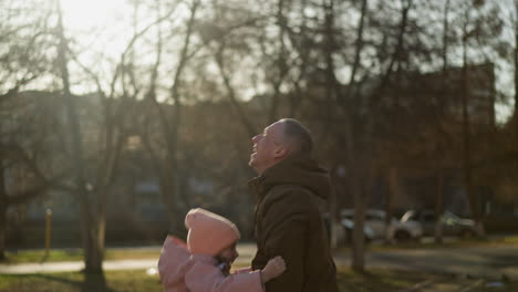 a little girl in a pink cap and jacket being joyfully thrown up in the air by a man wearing a brown jacket, as they both laugh together. the scene captures a playful and happy moment in a sunlit park