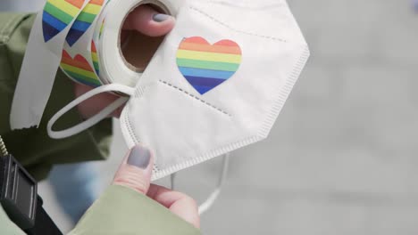 Cierre-De-Manos-Con-Uñas-Pintadas-Colocando-Una-Pegatina-En-Forma-De-Corazón-Lgbtq-Rainbo-En-Una-Máscara-Ffp2-Durante-Una-Demostración-De-Orgullo-En-Un-Día-Soleado