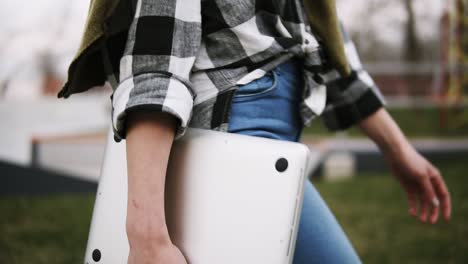 primer plano de una chica elegante en vaqueros camina por el parque con una computadora portátil en la mano. camina con confianza. vista lateral. sin rostro