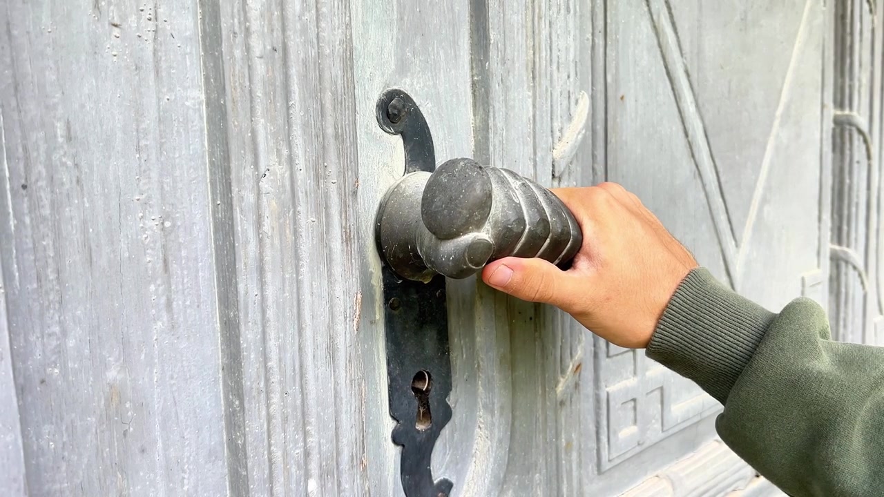 Close-up shot of a person's hand opening an old grey wooden door using a vintage fist-shaped metal handle. Ideal for historical documentaries, travel vlogs, and real estate ads.