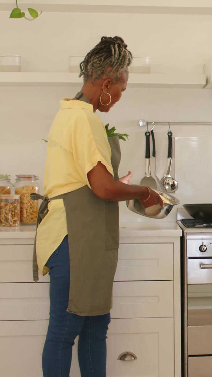 Vidéo verticale : Femme en tablier se penchant pour verser le contenu d'un bol en métal dans une poêle dans la cuisine, une main apparaissant.