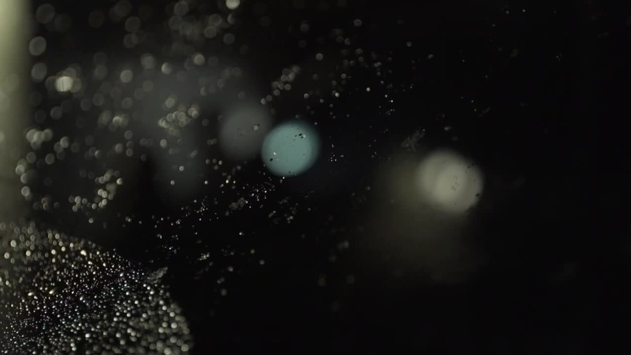 Snow melts onto night windshield with defocused lights in background